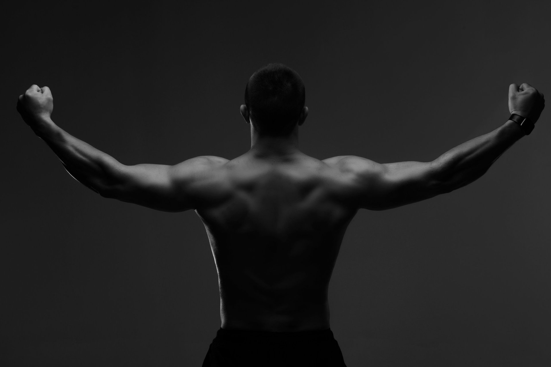 Man bodybuilder standing with arms raised, showing muscle definition in dark studio, black and white photo, back view